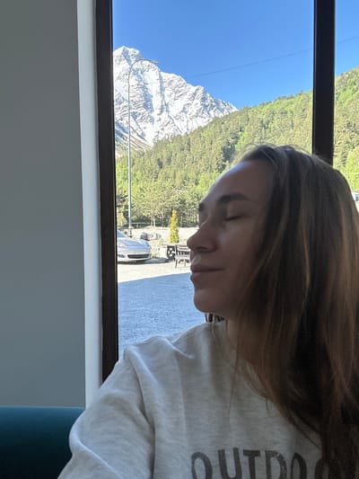 A young woman with closed eyes and long, brown hair is captured in a close-up shot. She is wearing a light-colored sweatshirt with the word "OUTDOOR" visible on it. She is seated, possibly in a cafe or residential setting, with a teal-colored couch visible in the lower left corner. Behind her, through a large window, a breathtaking mountain landscape unfolds. The majestic snow-capped peak of Mount Elbrus, a prominent feature of Terskol, Russia, dominates the background. Lush green forests cover the lower slopes of the mountains, and a clear blue sky stretches above. In the mid-ground, a silver car with a license plate is parked on a gravel surface, alongside outdoor seating and a small, ornamental tree. The overall scene depicts a serene moment of contemplation amidst a stunning natural environment.
