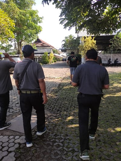 The image captures a scene from a cobblestone walkway in Gambiran, Indonesia (latitude: -8.434, longitude: 114.18). Four men are walking along the walkway, with the man closest to the camera wearing a dark gray shirt, black pants, and a blue baseball cap. The man second from the camera is wearing a dark gray shirt, black pants, and a black belt. The third man is wearing a dark blue shirt, black pants, and a black backpack. The fourth man is wearing a dark gray shirt, black pants, and a pair of white sneakers. The walkway is lined with trees, and the buildings in the background are white with a tiled roof. The sky is bright and clear.