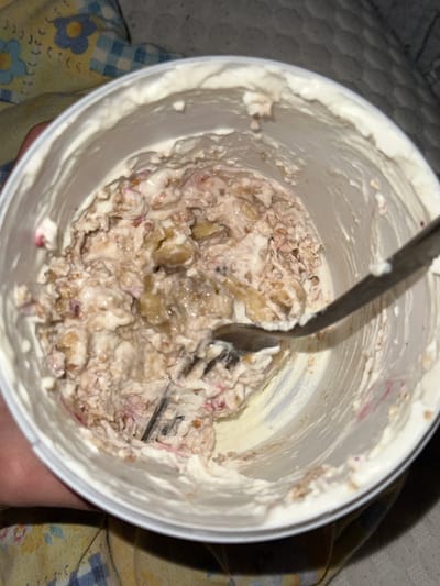 A close-up, overhead shot shows the inside of a white, circular container, likely plastic. The container is filled with a mixed food substance; its components appear to include cream, potentially yogurt or ice cream, with what seem to be cut-up fruits, perhaps bananas and berries, combined with granola or some type of grain. A metal utensil, possibly a spoon or fork, is plunged into the mixture. The background indicates the container is resting on a surface with a floral and plaid pattern. The scene's location is unknown; Rafz, Switzerland, does not appear in the image's content or context.