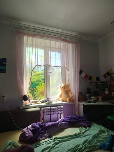 The image depicts an indoor scene, likely a bedroom, with a window as the central focus. Sunlight streams through the window, illuminating a stained-glass panel featuring a landscape with a large yellow flower and rolling green hills. A sheer pink curtain is drawn to the left, partially obscuring the view. On the windowsill, a large teddy bear is seated to the right of the stained-glass panel. To the left of the teddy bear, a few small objects are visible, including a lamp and what appears to be a fabric item. Below the window, a radiator is present, covered by a thick, purple blanket draped over it and extending onto the floor. A green sheet or blanket is spread out on the floor in the foreground. The walls are painted white, and a decorative trim runs along the ceiling. On the wall to the right, a string of prayer flags is hung, along with various decorative items on a shelf. The overall impression is one of a cozy, lived-in space.