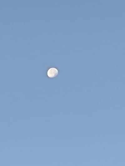The image is a photograph of the moon in a clear blue sky. The moon is partially illuminated and appears to be a waning crescent. The sky is a uniform light blue, with no clouds visible. The image is taken from a low angle, looking up at the moon. There is no indication of the city of Yuzhno-Sukhokumsk, Russia in the image.