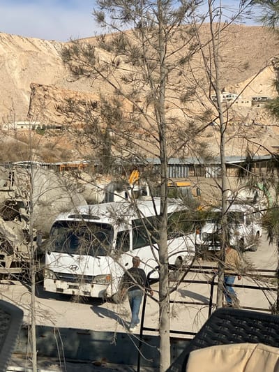 The image, taken from an elevated perspective, shows a street or yard scene in Dahiyat Harasta, Syria, on a sunny day. Bare tree branches and some green foliage heavily frame the foreground, partially obscuring the view.
In the midground, several white utility vehicles or trucks are visible. A prominent white truck, bearing a "Hyundai" logo on its front, is parked facing the viewer. A man in a dark top, blue jeans, and white sneakers stands next to the driver's side of this truck, appearing to interact with it. Further back, another man in a light-colored jacket and blue jeans is partially visible near a second white vehicle. A yellow construction excavator or similar machinery is also partly seen in the deeper midground. The ground is paved, likely asphalt or concrete, with strong shadows indicating bright daylight.
The background features a large, rocky, arid mountain or hill under a light blue sky with some white haze. A few light-colored buildings or structures are visible at the base of the mountain. On the far right foreground, a portion of a dark, woven outdoor chair or railing and a beige fabric object are visible. The overall scene depicts a daytime setting with men near utility vehicles, set against a characteristic mountainous landscape.