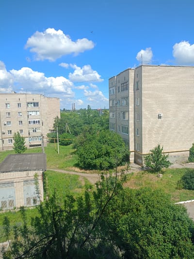 This is a daytime, outdoor, eye-level shot of a residential area in Gukovo, Russia, under a partly cloudy sky. The scene features two multi-story apartment buildings constructed of light-colored brick. The building on the left appears to be older, with visible balconies and many windows. The building on the right is similar in construction but has a more modern air conditioning unit mounted on its facade. In the foreground, a smaller, single-story brick building with a dark, flat roof is visible. This structure has two large grey doors, one labeled with "T1 250.00 A" and another with "T2 250.00 A", suggesting it might be a utility or garage building.
Lush green trees and vegetation surround the buildings, creating a sense of a well-established neighborhood. The grass is a vibrant green, indicating a warm season, likely late spring or summer. The sky is a bright blue, with fluffy white cumulus clouds scattered across it. A few small birds are visible in flight against the sky. In the distance, industrial smokestacks can be seen, hinting at the industrial nature of the region. The overall atmosphere is peaceful and serene, with no people or active events depicted. The sunlight suggests it is likely mid-morning or afternoon.