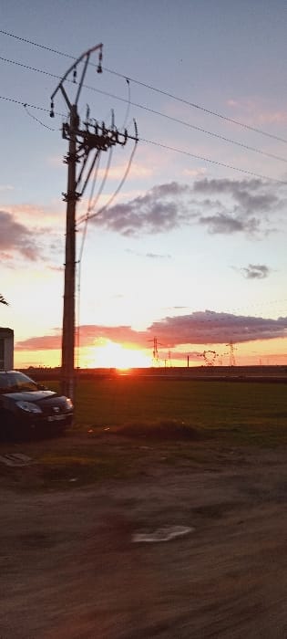 A sideways sunset scene showcases a telephone pole with power lines against a sky with clouds. Power pylons are visible in the distance. A field and a car appear along the horizon, adding depth to the shot. A road or dirt path occupies the foreground, contributing to the rural landscape.