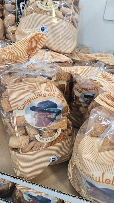 A close-up shot shows multiple transparent plastic bags filled with light brown cookies. The bags are adorned with an orange and white striped pattern. A circular label is visible on the front of one bag, featuring a profile image of an elderly man wearing a hat, alongside text in French. Another bag displays the text "Cuggiulielle de Zilia". The cookies themselves are small, irregularly shaped, and appear to have a coarse texture. The bags are packed tightly together, suggesting a retail or bulk storage setting. The image is taken from a slightly elevated angle.