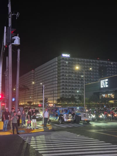 At night in Pasay, Philippines, a crowd of people cross a pedestrian zebra stripe at a crosswalk in front of a large building. The building features an “SMDC” sign on its left side and a “MET LIVE” sign above. On the street next to the crosswalk, many parked motorbikes and tricycles are aligned, with one tricycle being brightly colored. Street lights and traffic lights are present. The sky is dark.