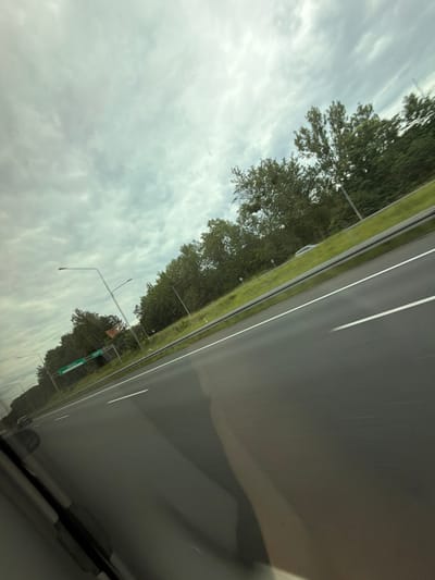 The image captures a view from a vehicle traveling on a highway in Ruda Śląska, Poland, on a cloudy day. The highway is paved and has white lane markings. On the right side of the highway, there is a grassy embankment lined with trees. There are also streetlights visible. A green road sign with white lettering is present on the left side, indicating a direction or destination. Another vehicle is visible in the distance on the left side of the highway. The sky is overcast with gray clouds. The perspective is angled, suggesting the photo was taken from a moving car.