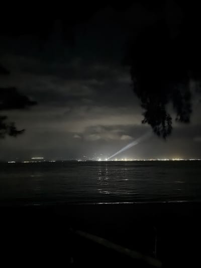 A nighttime, low-light view from a dark foreground, possibly a beach, looking out over a body of water towards a distant city skyline. The water is dark and appears to have gentle ripples reflecting faint lights. The city skyline is a line of scattered lights, some appearing as clusters and others as individual points. A prominent, bright beam of light originates from the city and shoots upwards into the cloudy sky. The sky is dark with patches of lighter clouds, illuminated by the distant city lights. A dark silhouette of foliage hangs from the upper right corner of the frame. The scene appears to be a coastal area in Melaya, Indonesia, likely captured during the evening or night.