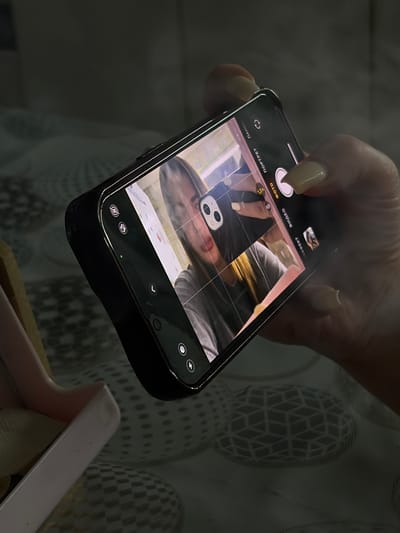 A person's hand holds a black smartphone, capturing an image of a woman wearing a dark shirt, who appears to be taking a selfie with another phone with two visible cameras on the back. The background features fabric with a geometric pattern of ovoid shapes. The phone being used to take the photo is above what appears to be a partially visible box. No evidence suggests this scene is located in Port-Katon, Russia.