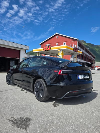 The image shows a black Tesla parked in front of a red building with a yellow canopy, likely a gas station. The building has large "5" logos visible. The Tesla has a spoiler on the trunk and a Norwegian license plate that reads "ER 26329". In the background, there are mountains and a bright blue sky with some white clouds. Two birds are visible flying in the sky. The ground appears to be asphalt, and there are some dark stains on it. The scene suggests a sunny day, possibly in the late morning or afternoon, given the lighting and shadows. The location appears to be a somewhat rural or scenic area, as indicated by the mountains. The overall mood is peaceful and sunny.