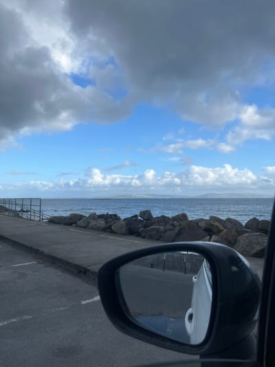 The image, taken from inside a car, shows a coastal scene in Galway, Ireland. A car's side mirror is prominently featured, reflecting a section of pavement and another car parked behind. Outside the car window, a concrete pier runs along the water's edge, protected by a barrier of large rocks. The water stretches to a distant coastline under a sky with alternating patches of blue and heavy grey clouds.