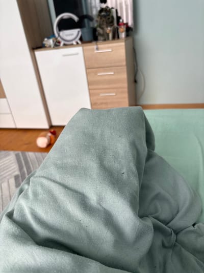 The scene depicts an interior residential space in Nadarevo, Bulgaria. In the immediate foreground, a substantial amount of crumpled, light green fabric, identified as a blanket or duvet, dominates the lower half of the frame, showing small dark particulate matter on its surface. The fabric extends upwards, suggesting it covers an unseen subject or object. To the left, a section of light wooden flooring is visible, partially covered by a grey and white geometric patterned rug. An upright white and light wooden cabinet or wardrobe unit is positioned against a light blue-green wall. The unit consists of a taller white section on the left and a shorter light wood section with three drawers on the right. On top of the wood section, a white circular ring light stands beside a multi-colored figurine, possibly a cat, and other indistinct objects, including a small red and white container. A light brown cylindrical object with a red top, possibly a bottle, rests horizontally on the wooden floor to the left of the blanket. No individuals are visible.