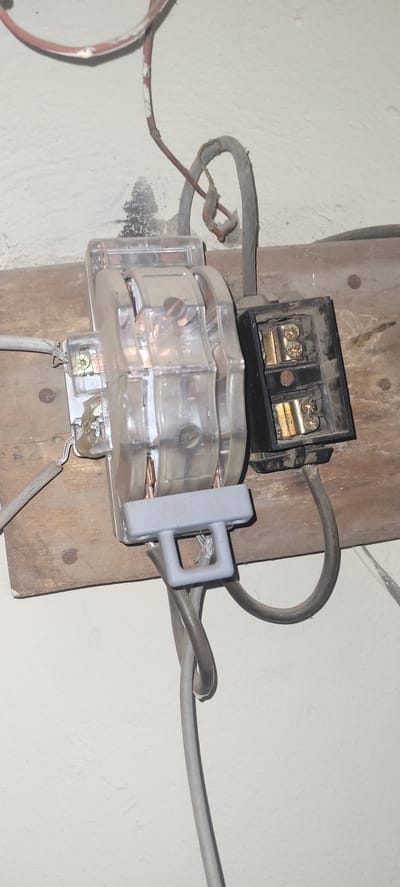 The image depicts an electrical connection mounted on a wooden board against a textured wall. A clear plastic electrical component with copper conductors is centrally located, with several wires connected to it. To the right of this, a black electrical junction box with brass terminals is visible. Various electrical cables, some appearing worn or damaged, run to and from these components. The overall setting suggests an electrical installation, likely in a residential or commercial space, with evidence of potential wear and tear. The specific location within Jalingo, Nigeria, cannot be determined from the visual information provided.