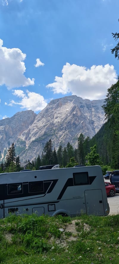 This is a medium shot, taken outdoors in a vertical orientation, of a motorhome parked on a dirt road next to a grassy slope. In the background, a large, rocky mountain dominates the scene, with a bright blue sky and white clouds above. The mountain has visible striations and appears rugged and imposing.

The motorhome is modern and grey, with dark accents and several windows. Text on the side of the motorhome reads "Sonic SUITE". Several cars are parked behind the motorhome. The grassy area in the foreground is overgrown with wildflowers, suggesting a natural, undeveloped setting. Evergreen trees are scattered throughout the scene, lining the base of the mountain and visible behind the motorhome.

The time of day appears to be daytime, likely mid-morning or afternoon, given the brightness of the sky and the shadows cast. The weather seems clear and sunny, with fluffy white clouds indicating fair conditions. The combination of the motorhome, the parked cars, and the stunning mountain landscape suggests this is a popular tourist area, possibly a campsite or a scenic viewpoint. The overall mood is peaceful and serene, capturing a moment of travel or vacation in a beautiful natural environment.