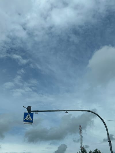 An upward-angle, medium shot captures a cloudy sky over a cityscape. Dominating the left side of the frame is a pedestrian crossing sign, a blue square with a white triangle in the center, featuring a white silhouette of a person walking. The sign is mounted on a horizontal pole, which curves downwards on the right. A tall, lattice telecommunications tower with red and white accents is visible in the background on the right, partially obscured by trees. The environment appears to be a public road area within the Плешчаніцкі сельскі Савет, Belarus. The sky is filled with a mixture of white and gray clouds, suggesting an overcast day.