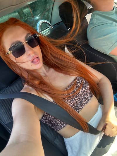 The image shows two people in a car. One person, likely a woman with long red hair, is lying down and looking at the camera with sunglasses on and her tongue sticking out. She is wearing a leopard-print bikini top and white shorts. A seatbelt is across her body. Another person, whose body is partially visible on the left of the frame, is wearing a light green t-shirt. The car appears to be parked, with a metallic loop visible on the dashboard. The scene suggests a casual outing, possibly on a sunny day. The location is identified as Golubovići, Montenegro.