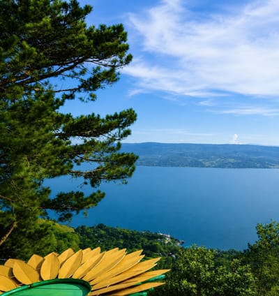 A high-angle, full-shot captures a panoramic view of a vast, calm, deep blue lake bordered by verdant, tree-covered hills and mountains. The sky is a vibrant blue with scattered white clouds. In the foreground, a large, stylized yellow flower sculpture with green accents is partially visible on the left, adding a decorative element to the natural landscape. The overall scene is serene and picturesque. The location is identified as Parik Sabungan, Indonesia.
