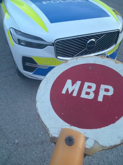 The image shows a close-up, slightly overhead view of the front section of a police car positioned next to a circular handheld sign.
The police car is a Volvo, with its emblem visible on the front grille. Its livery is white, accented with prominent blue sections and fluorescent yellow (or lime green) stripes and patterns. The word "POLICE" is partially visible in white text on the blue section of the car's front.
To the right of the police car is a circular sign featuring a red center and a white outer ring. Bold white capital letters "MBP" are displayed across the red area of the sign. The sign appears worn, with visible chips and scuff marks on its edges and surface. It is attached to a yellow handle or pole, with a metal screw securing it.
The scene is set outdoors on what appears to be a grey, paved surface, likely asphalt. The lighting indicates it is daytime. The combination of the police vehicle and the "MBP" sign (often associated with police or interior ministry functions, particularly in a Bulgarian context) suggests an activity related to law enforcement, such as a checkpoint or traffic control. The location context is Smyadovo, Bulgaria.