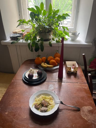 A medium, overhead shot shows a wooden table next to a window with a plant in it. On the table, a plate of cooked cabbage with meat sits on the right side. Adjacent to it, closer to the window, is a plate with two dark, round objects and a small, light-colored object. A bowl of oranges and a banana are situated near a long, red object and a small, patterned box. The scene appears to be indoors. It is not possible to determine if the location is in Перовское сельское поселение, Ukraine based on the visual content.