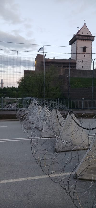 The image depicts a segment of road barricaded with razor wire in Ivangorod, Russia. In the background, the Ivangorod Fortress is visible against a cloudy sky. A metal fence runs parallel to the road, and the Estonian flag is seen. There is no visible human activity.