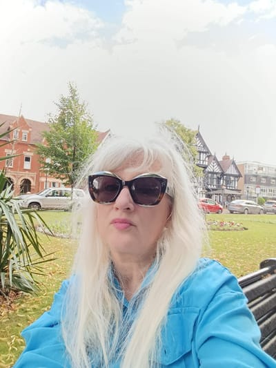 A woman with blonde hair and sunglasses is taking a selfie in a park. She is wearing a blue shirt. In the background, there is a brick building and a Tudor-style building. There are also cars parked on a street behind the park. The location appears to be Harpur Square in Bedford, United Kingdom, as suggested by the presence of the distinctive architecture of The Old Town Hall and the adjacent buildings. The sky is cloudy.