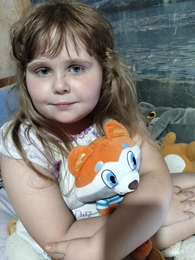 The photograph, taken indoors in the city of Shchastia, Ukraine, shows a young Caucasian girl with blonde hair and blue eyes. She is holding a stuffed animal, a cartoon fox character, with orange and white fur and blue eyes. The girl is wearing a white dress with purple trim. In the background, there are other stuffed animals, including a large gray elephant and a brown dog-like stuffed animal. The overall environment appears to be a bedroom. A small scratch is visible on the girl's forehead.