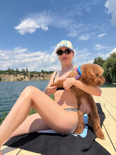 The media file shows a person holding a small, fluffy, light brown dog. The person is wearing a black swimsuit, sunglasses, and a white baseball cap with a patch on the front. The dog has a blue collar with some patterned accents. They are sitting outdoors on a wooden deck next to a body of water. In the background, there is a rocky hillside covered in green trees and foliage. The sky is blue with scattered white clouds, suggesting it is daytime and the weather is pleasant. The overall scene appears to be a relaxed, sunny day spent by the water. The word "VICTORIA'S" is visible on the strap of the swimsuit. The location context is Novomykolaivka, Ukraine.