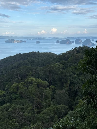 An aerial landscape view captures a lush, dense forest in the foreground, sloping downwards towards a calm, blue ocean. Numerous verdant islands of varying sizes dot the ocean's surface, appearing as dark shapes against the water. In the distance, layers of hazy blue mountains form the horizon, beneath a sky filled with a mix of white clouds and clear blue patches. The overall scene depicts a tropical, natural environment. The location is identified as Ban Din Daeng Nui, Thailand. There are no people or animals visible in the image.