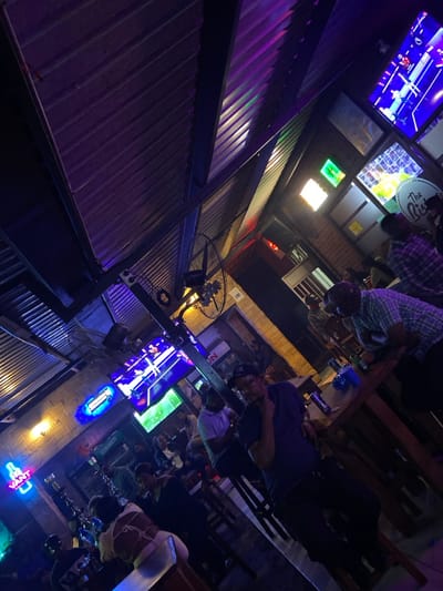 The image shows an outdoor bar area with a corrugated metal roof, located at coordinates -6.157 latitude and 35.724 longitude in Dodoma, Tanzania. The bar is dimly lit with purple and blue lights. There are several patrons seated at tables, and some are standing and talking. Several patrons have drinks in their hands. There is a large screen showing a sporting event in the background, and a sign that says "The Ritz" is visible on the wall. In the foreground, a man wearing a black shirt and a black baseball cap is sitting at a table. He is looking down at his phone, and his hand is resting on his chin. In the background, a man wearing a plaid shirt is talking to someone who is out of frame. The overall atmosphere is relaxed and casual.