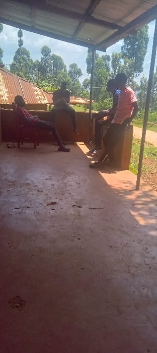 The image depicts an outdoor, partially covered area within Getenga ward, Kenya. Five individuals, appearing to be males, are present. One individual on the far left is seated on a red plastic chair, wearing a dark red shirt and dark trousers, with legs extended. Four additional individuals are seated on a low, earth-toned wall or platform to the right, facing generally towards the left or center of the frame. One of these individuals wears a light-colored, possibly pink, short-sleeved shirt. The ground consists of a concrete slab, showing wear and discoloration, partially covered by shadow. Above, a corrugated metal roof provides shelter, supported by visible structural elements. In the mid-ground, behind the seated individuals, structures with rusted corrugated iron roofs are visible amidst dense green foliage and tall trees. A dirt path or road is present on the far right of the scene. The sky is partly cloudy.