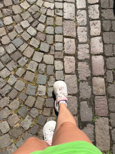 An overhead, eye-level shot captures a person's legs and feet from the knee down, walking on a cobblestone street. The person is wearing white Crocs with pink accents, white socks, and neon green shorts. The cobblestones transition from small, irregular grey stones to larger, rectangular pink and grey stones, and finally to dark grey asphalt pavers. Green weeds are growing between the stones. The scene is brightly lit by sunlight, casting shadows. The location is identified as Telgte, Germany.