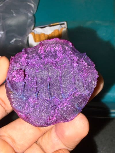 A close-up, overhead shot shows a person's hand holding a rough, purple and blue mineral specimen. The mineral is irregularly shaped, with a textured, crystalline surface. In the blurred background, to the left, is a crumpled clear plastic bag. To the right of the bag, a small, white box contains several elongated, orange-colored objects, possibly food items or pills. The background is a dark, teal-colored surface with some shadows. The lighting is dim, and the focus is on the mineral specimen and the hand holding it. The image does not contain any contextual information that would allow for the determination of its location in Tupah, Indonesia.