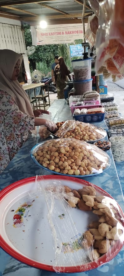 The image shows a street food stall in Pandrahkandeh, Indonesia. A woman wearing a floral-patterned dress and a hijab is selling Indonesian snacks. The food is displayed on a blue table covered with a plastic sheet. There are several trays of snacks, including small, round, light brown pastries covered in plastic wrap. There are also other snacks in baskets and plastic containers. A banner with the words "Hari Kartini & Baksos IBI" hangs in the background. A woman and a man, likely customers, are standing behind the stall. A fan and a motorcycle are visible in the background.