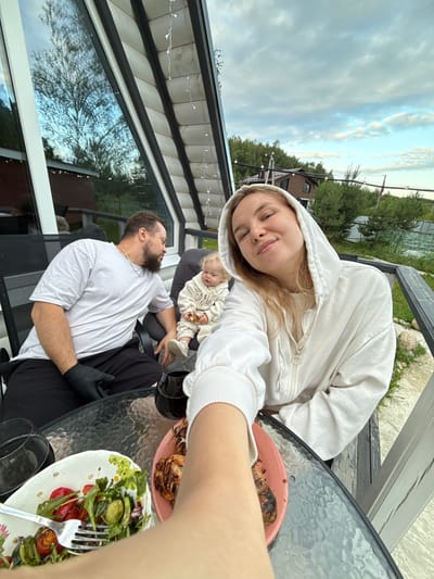 In Ryzhkovo, Russia, on a patio with a view of trees and houses under a cloudy sky, a family enjoys a meal. A woman in a white hooded jacket is taking a selfie. Next to her, a baby in a cream-colored onesie sits on a chair, holding a cracker. A man in a white t-shirt and black pants with black gloves sits on a chair, looking at the baby. A glass table holds two glasses of red wine, a salad in a white bowl, and grilled chicken on a pink plate. The patio has a railing and a sloped roof with string lights.