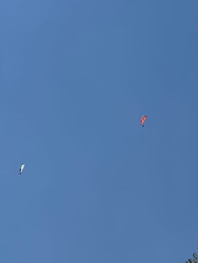 The media file shows a clear blue sky with two kites flying. One kite is orange and has the word "Gazer" visible on it. The other kite is white and green. The image appears to be taken during the day, with good weather conditions for kite flying. There are no people or obvious location cues in the image, other than the provided location context of Čučuci, Montenegro. The activity taking place is kite flying.