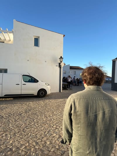 A man stands on a stone-paved street in Vila Real de Santo António, Portugal, his back to the camera. To the left, a white building with a window and a parked white van is visible. A group of people are seated at tables in the background. The sky is blue.