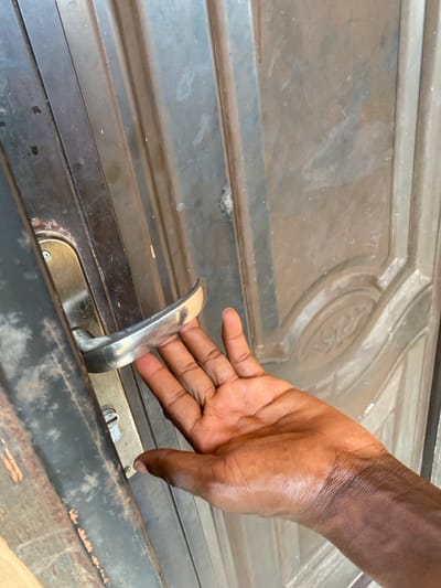 A close-up shot focuses on a person's hand reaching for the handle of a weathered, dark-colored metal door. The hand, with its dark skin and visible creases, is positioned to grasp the curved, silver door handle. The door itself appears to be made of metal and has a raised, decorative panel with a circular design in the upper right portion of the frame. The surface of the door is dusty and shows signs of age and use, with subtle scratches and smudges. Below the handle, a lock mechanism is visible, with a screw head and a keyhole. The scene appears to be outdoors, given the texture and wear of the door, and the lighting suggests it could be daytime. The image captures a simple, everyday action of opening or closing a door. The setting is identified as Bulamare, Nigeria. There is no visible text in the image.