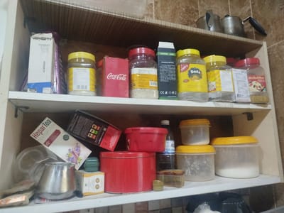 The image shows a cluttered shelf in a kitchen or pantry, likely in Sabunchu, Azerbaijan. Various food items and containers are visible, suggesting a domestic setting. On the top shelf, two metal kettles sit next to a shelf lined with boxes and jars. The middle shelf is more densely packed, featuring jars of food with yellow lids, a Coca-Cola box, and a "Camping Lamp" box. The bottom shelf holds a variety of containers, including a "Hepar Tea" box, a red tin, and several plastic jars with yellow lids. Some of the jars appear to contain white powdery substances. The overall impression is one of a well-used kitchen storage area. Visible text includes brand names like "Coca-Cola," "Besta," and "Hepar Tea," along with product descriptions and manufacturing information. The lighting suggests it might be daytime, but the interior setting doesn't provide explicit clues about the weather or time of day. No people are visible, and the scene evokes a sense of everyday domestic life.