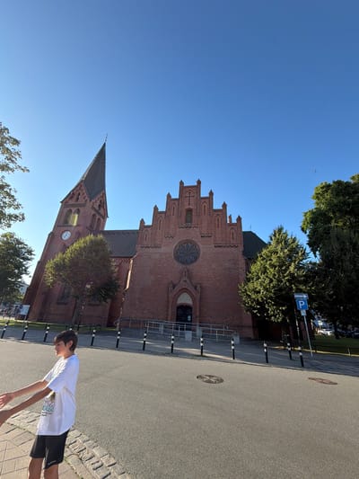 In Rostock, Germany, a boy in a white t-shirt and black shorts walks past a brick church with a tall spire and a rose window. The sky is clear and blue. The street in front of the church is paved, and there are bollards along the sidewalk. A parking sign is visible near a tree.