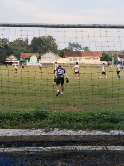 A low-angle outdoor shot, taken through a soccer goal net, shows a soccer field with players during what appears to be a training session or a relaxed game. Several players are visible on the field, wearing different colored jerseys with numbers on their backs, including "50" and "22." The players are spread out across the field, some standing, some walking.
In the background, there are buildings. The building with the sign “TK DESA BOTOK” is on the left side of the image and another building with the sign “SD NEGERI 01 BOTOK KEC. KERJO” is on the right. These signs provide location information: Botok, Indonesia. The background also includes trees and greenery.
The foreground is dominated by the soccer net, which is slightly out of focus, and some weeds and plants.
