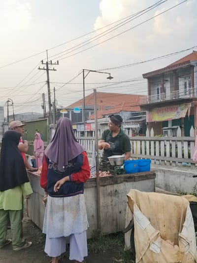 In Barengkrajan, Indonesia, an outdoor market scene unfolds under a hazy sky. In the foreground, a vendor wearing a dark green shirt and apron stands behind a stall laden with raw meat. He is gesturing while speaking to a customer, a woman in a purple hijab and a red and blue jacket. To the left, two other individuals are present: one woman in a black hijab and light green attire, and another person partially visible. The stall itself features a scale, two blue containers, and a large piece of tan fabric covering something in the lower right of the frame. In the background, a white picket fence separates the market area from buildings. One building displays a yellow banner with Indonesian text that reads "TOKO NAIKILA," indicating a store. Another building has a sign with "MB KOMUNIKASI." Electrical poles and numerous wires crisscross the sky, typical of an urban Indonesian setting. The overall atmosphere suggests a bustling commercial exchange at a local marketplace.