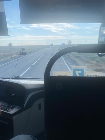 The image depicts a view from inside a vehicle, likely a bus or truck, looking out onto a highway. The perspective suggests the vehicle is in motion. The sky is visible, filled with scattered white clouds against a bright blue backdrop, indicating daylight. The highway stretches out ahead, marked with white dashed lines, and appears to be a two-lane road with a median. Signs are visible along the roadside, including a circular sign with the number "50" inside a red border, likely a speed limit sign, and a blue directional sign with a white arrow pointing left, indicating a turn. Another sign with text is partially visible, with "HAY" and "ENDUNG" being legible, potentially indicating place names or route information.

The interior of the vehicle is partially visible, showing a dashboard with various controls and a steering wheel. A camera is mounted on the dashboard. The overall setting is a rural or semi-urban environment in Kazakhstan, given the location context of Қосшағыл ауылдық округі. The mood appears neutral, focusing on the journey and the road ahead. The time of day is daytime, and the weather is clear with some cloud cover.