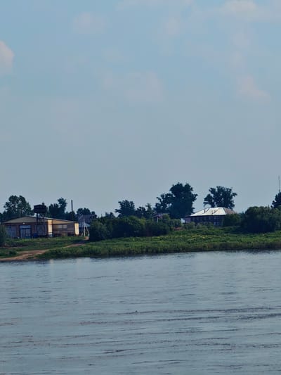 The image depicts a wide, calm body of water in the foreground, likely a river, with ripples on its surface. In the background, a green, grassy embankment is lined with trees. Several buildings are visible on the embankment. To the left, there is a long, rectangular building with a light-colored facade and a darker roof. Adjacent to it is a metal structure with a tank on top. Further to the right, there is a larger, two-story building with a dark, sloped roof and visible windows. The sky above is a light blue with a few scattered white clouds. The scene appears to be a rural or semi-rural area, possibly on the outskirts of Yeniseysk, Russia, given the characteristic landscape and building styles often found in the region. There are no people or animals visible in the frame. The time of day appears to be daytime, with diffused sunlight.