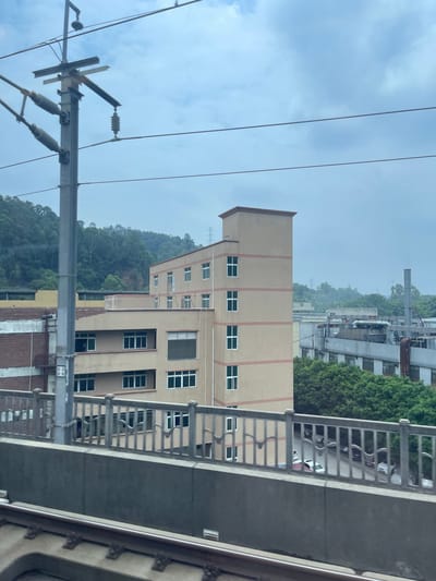 The image shows a view of buildings and power lines against a cloudy sky. A light brown, multi-story building with numerous windows is prominently featured in the foreground, with a darker concrete structure and metal railing to its right. Beyond this building, more industrial-looking structures and trees are visible on a hillside, with a power transmission tower in the distance. The scene appears to be outdoors, possibly from a window or balcony. The overall impression is of an urban or industrial setting with natural elements. The lighting suggests it is daytime, but the overcast sky indicates potentially cloudy or rainy weather. There is no clear activity or event taking place. Visible text on a small white sign on a pole is not legible. The orientation of the image is rotated 90 degrees counter-clockwise.