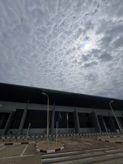 The image displays the exterior of an airport terminal building, identified by its green signage as "Sule Lamido International Airport," located in Kwi, Nigeria. The building exhibits a modern architectural design with a dark, flat roofline, grey structural elements featuring slanted panels, and dark, reflective glass windows. Two flagpoles are visible in front of the building, one flying a green and white flag, likely the national flag of Nigeria.

The sky is heavily overcast with dense altocumulus clouds, characterized by numerous small, patchy, white and grey cloudlets arranged in regular patterns, covering the majority of the visible sky. A bright, diffused patch of sunlight is visible through a thinner section of the clouds in the upper right quadrant of the image.

In the foreground, a paved area, likely a drop-off or parking zone, is present. It features white road markings, including lines delineating parking spaces or lanes, and a pedestrian crossing with broad black and white stripes. Black and white striped curbs and bollards delineate the edges of the paved areas and a small, sandy island where one of the lampposts is situated. Two beige lampposts, one prominently in the mid-ground and another further to the right, extend upwards towards the sky. No individuals, vehicles, or discernible activities are present in the frame. The overall impression is one of a quiet, modern airport facility under a cloudy daytime sky.