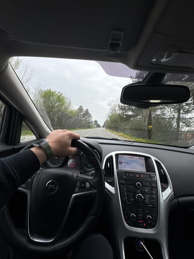 The image shows an interior view from the driver's seat of a car, traveling on a road lined with trees and vegetation on an overcast day. The driver's hand is visible on the steering wheel, wearing a smartwatch with a green band. The car's dashboard features a radio display tuned to 108.0 MHz, along with various buttons and controls. A black car air freshener hangs from the rearview mirror. The odometer reads 252259 km. I am unable to confirm its location in Musachevo, Bulgaria.