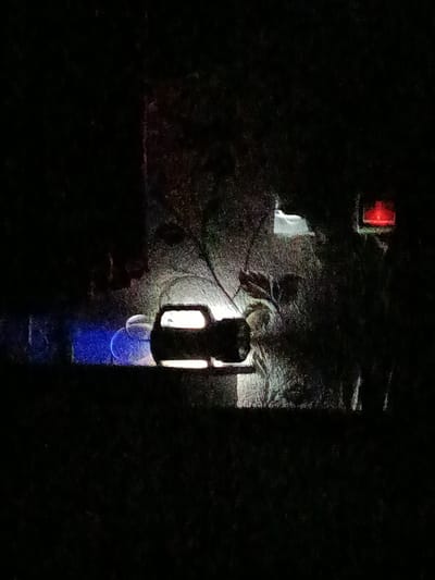 The media file depicts a nighttime scene, possibly in Ӯртабуз, Tajikistan, characterized by low light conditions and a slightly grainy texture. The central focus is a dark, somewhat blurry object that appears to be a vehicle, possibly a pickup truck, with its headlights illuminated, casting a bright white light forward. To the left of the vehicle, there are two glowing blue circular objects, suggesting some form of lighting or perhaps decorative elements.

Behind the vehicle, the background is a mix of dark foliage and faint, colorful lights. A white, rectangular object is visible above and behind the vehicle, possibly a sign or a structure. To the far right, a red, rectangular light fixture is prominent, casting a distinct red glow. The overall impression is of an outdoor setting at night, with artificial lights creating points of interest in the darkness. There is no visible text in the image. The scene is quiet and still, with no clear indication of people or specific activities, creating a sense of mystery or nighttime ambiance.
