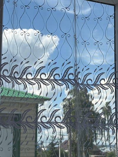 The image shows a sheer curtain with an intricate, dark grey embroidered floral pattern, viewed from indoors. Through the curtain, a bright blue sky with scattered white clouds is visible. The curtain is framed by a white window frame on the right side.
Below the sky, the pattern of the curtain changes to a repeating design of swirling, fern-like fronds. Through this lower portion of the curtain, a building with a light-colored, horizontal siding and a green roof is visible. The building appears to be one or two stories high. Behind the building, trees with green foliage can be seen, suggesting an outdoor environment.
The scene is likely set in a residential area. Given the specific location of Alexeyevskoye, Russia, this could be a view from a home, an apartment building, or a public building with a window overlooking a yard or street. The light suggests it is daytime, likely sunny given the brightness of the sky and clouds. There are no people or animals clearly depicted in the image.