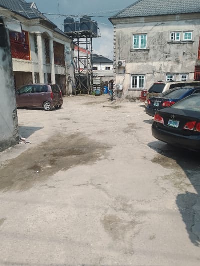 This image depicts an unpaved, irregular ground area, likely a courtyard or parking lot, in Mgbuoshimini, Nigeria, surrounded by multi-story buildings under an overcast sky.

The ground is a light-colored, worn surface, possibly compacted earth or old concrete, showing numerous dark, irregular stains consistent with fluid spills or moisture.

On the left, a two-story building with a light-colored facade features dark roof tiles, multiple windows with security bars, and a prominent ground-floor entry with columns. A maroon compact car or minivan is parked facing slightly left, its rear visible. A partial, weathered wall section is present in the extreme foreground on the left.

In the background-center, a black metal framework supports three large, black plastic water storage tanks. Blue barrels are visible at the base of this structure. Further structures with dark roofs are visible behind the water tower.

On the right, a two-story building with a heavily stained and discolored light-colored facade displays multiple windows with white frames, including one partially open on the upper floor. Two outdoor air conditioning units are mounted on this building's exterior wall. A row of vehicles is parked along the right side: a black Honda sedan (license plate KNG-774AF) is closest to the viewer, followed by a partially visible blue sedan (license plate appearing to be AAA-234), and a gold or tan SUV.

No individuals or animals are present. No specific actions or interactions are depicted.