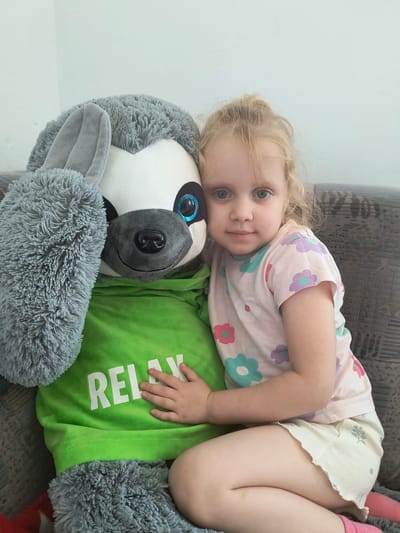 A young girl with blonde hair pulled up in a bun sits on a couch next to a large stuffed sloth. The sloth is wearing a green hooded sweatshirt with the word "RELAX" printed in white letters. The girl is wearing a pink t-shirt with a floral pattern and cream-colored shorts. She has her arm around the sloth. The background is a plain wall and the couch upholstery is textured. The lighting appears to be natural, coming from a window or overhead light. This image was taken indoors.